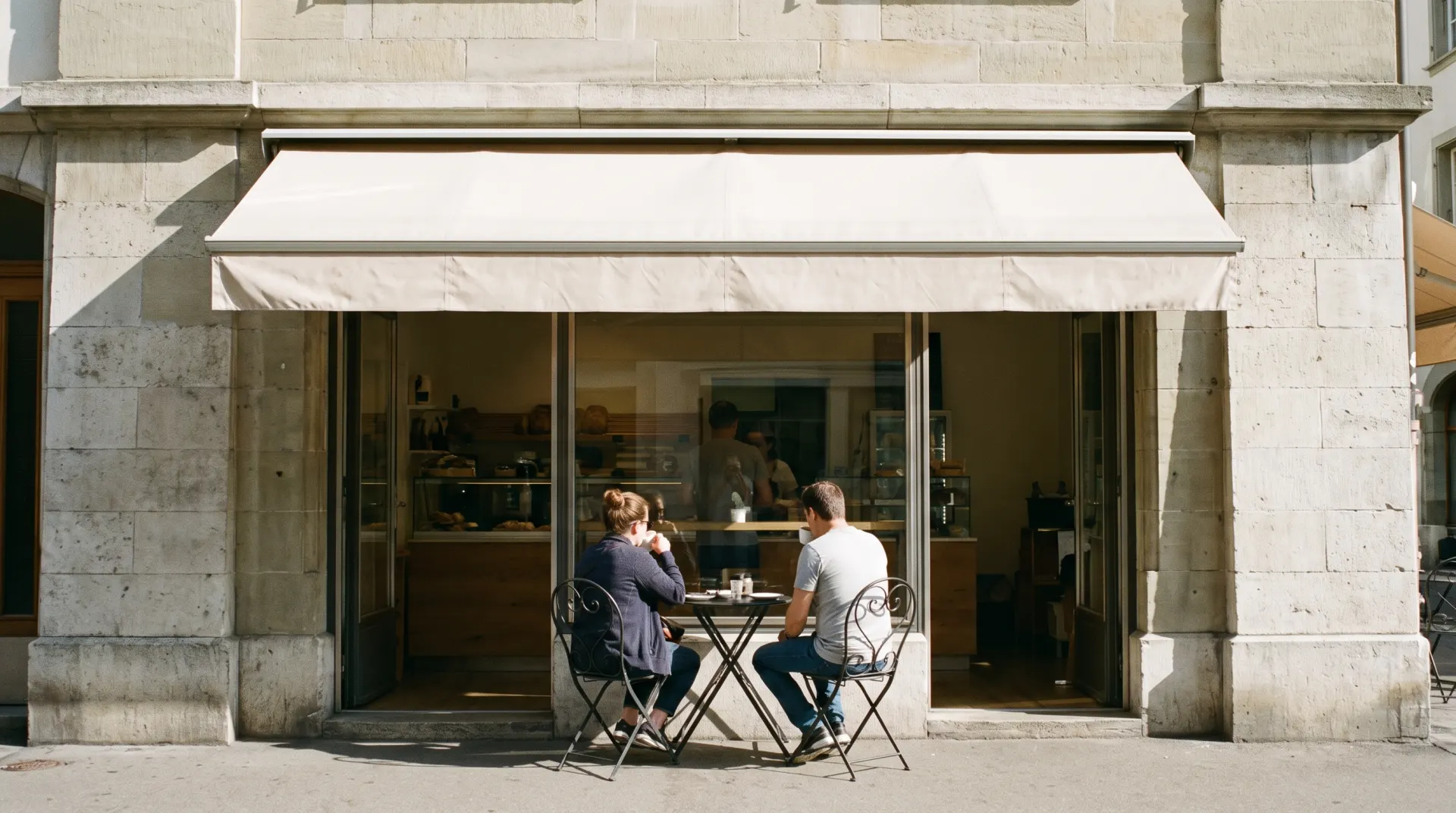 Stone front of a bakery café seen from the pavement, two customers on the terrace, the owner behind the window, in daylight